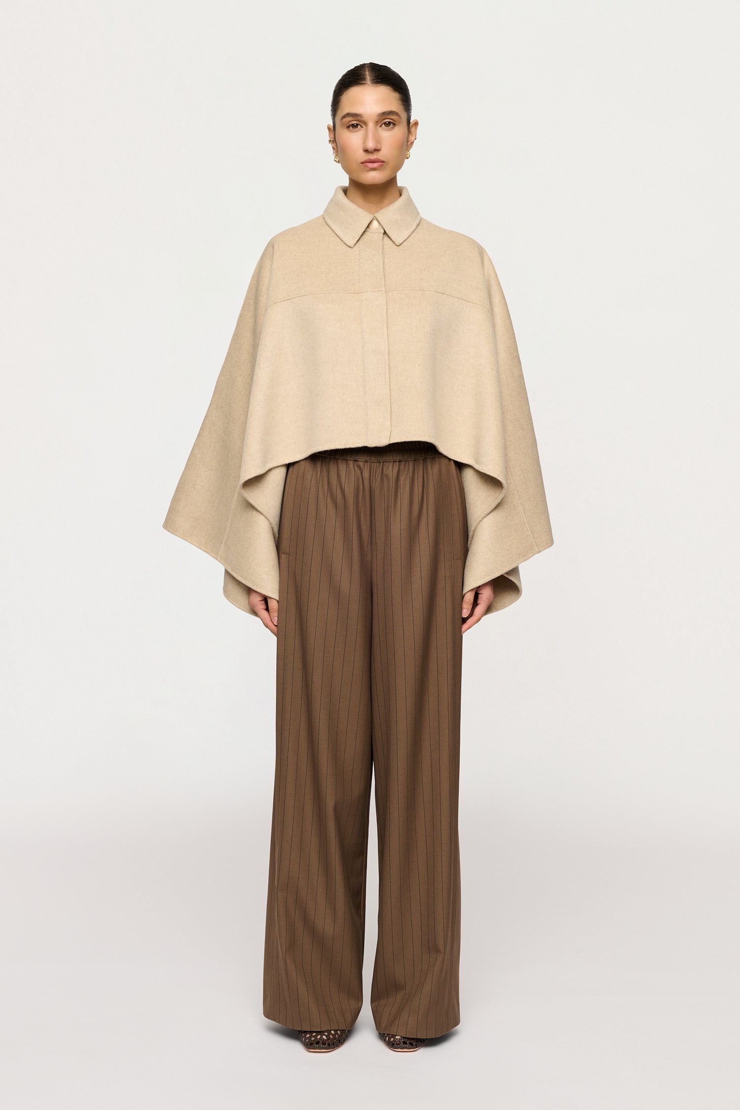 Person wearing a beige cape over a brown outfit on a white background