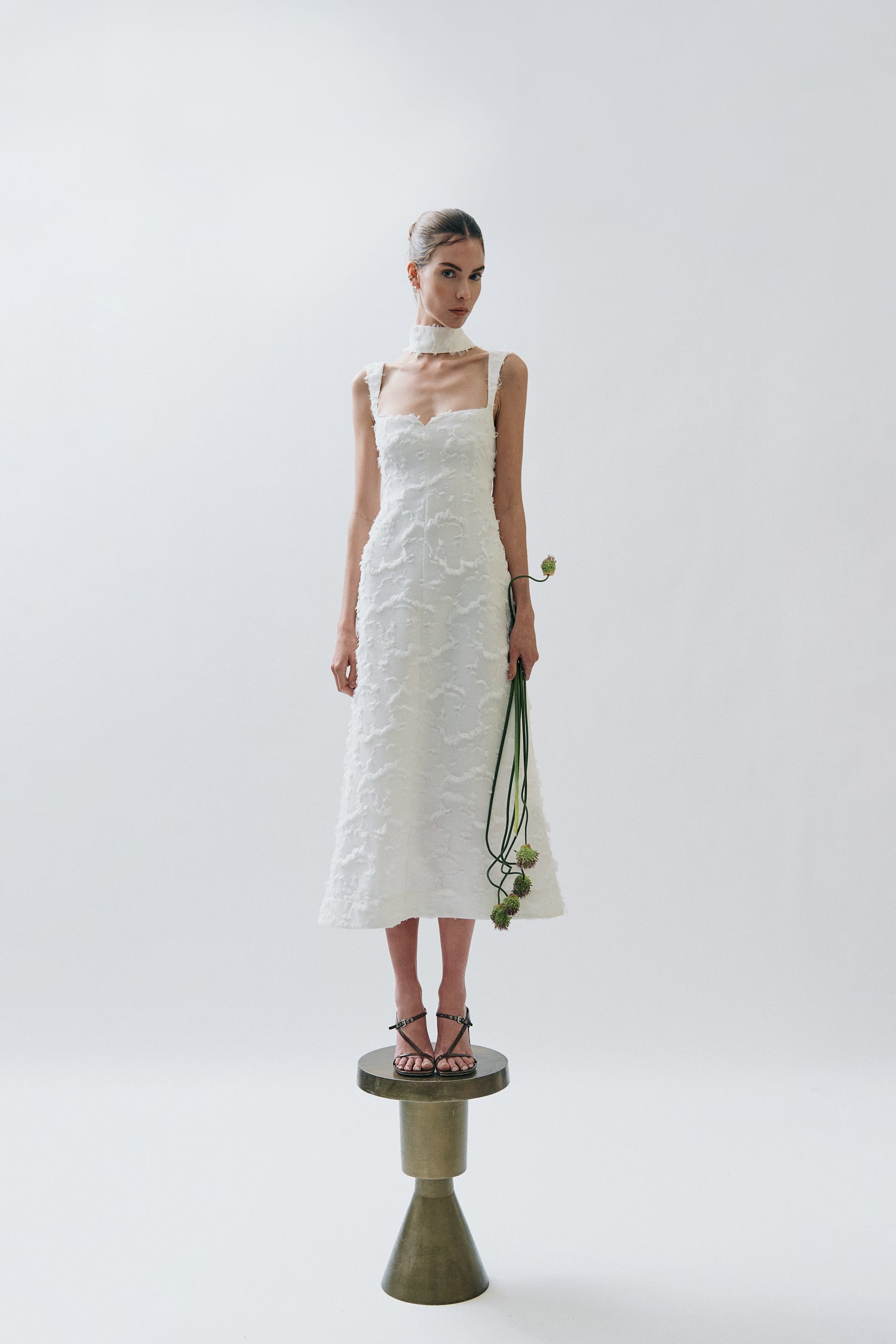Woman in a white Clea dress with floral textured details holding flowers on a pedestal against a white background