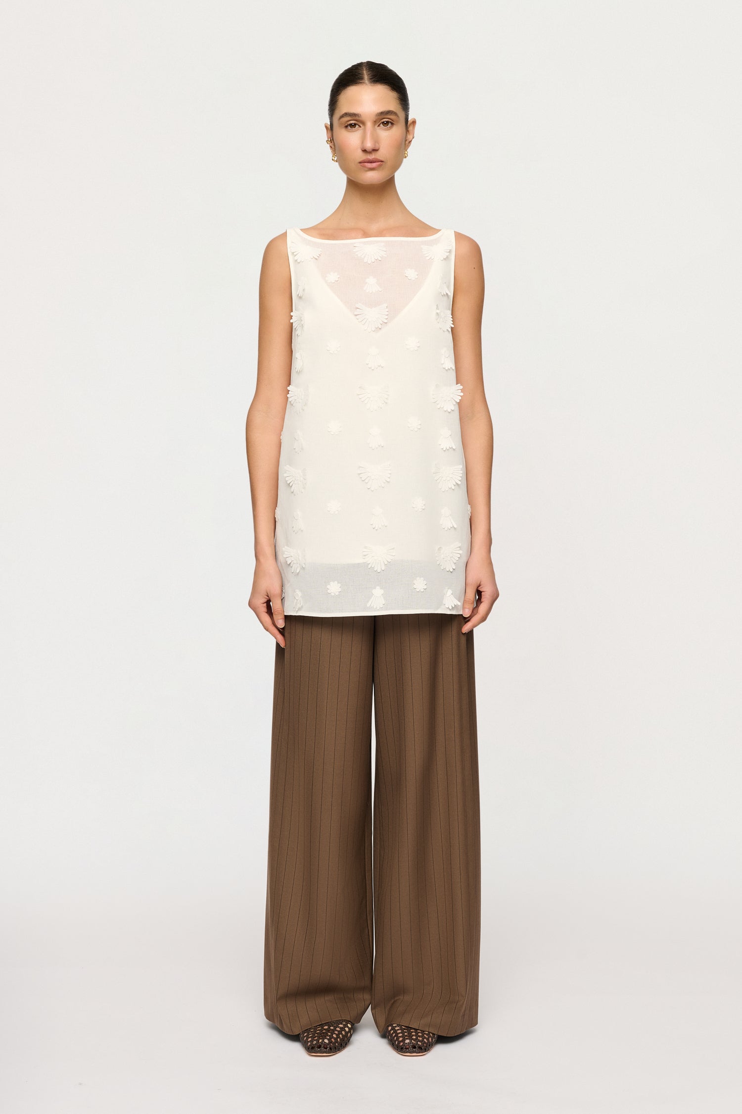 Woman wearing a white sleeveless top with a geometric pattern and brown wide-leg pants on a white background