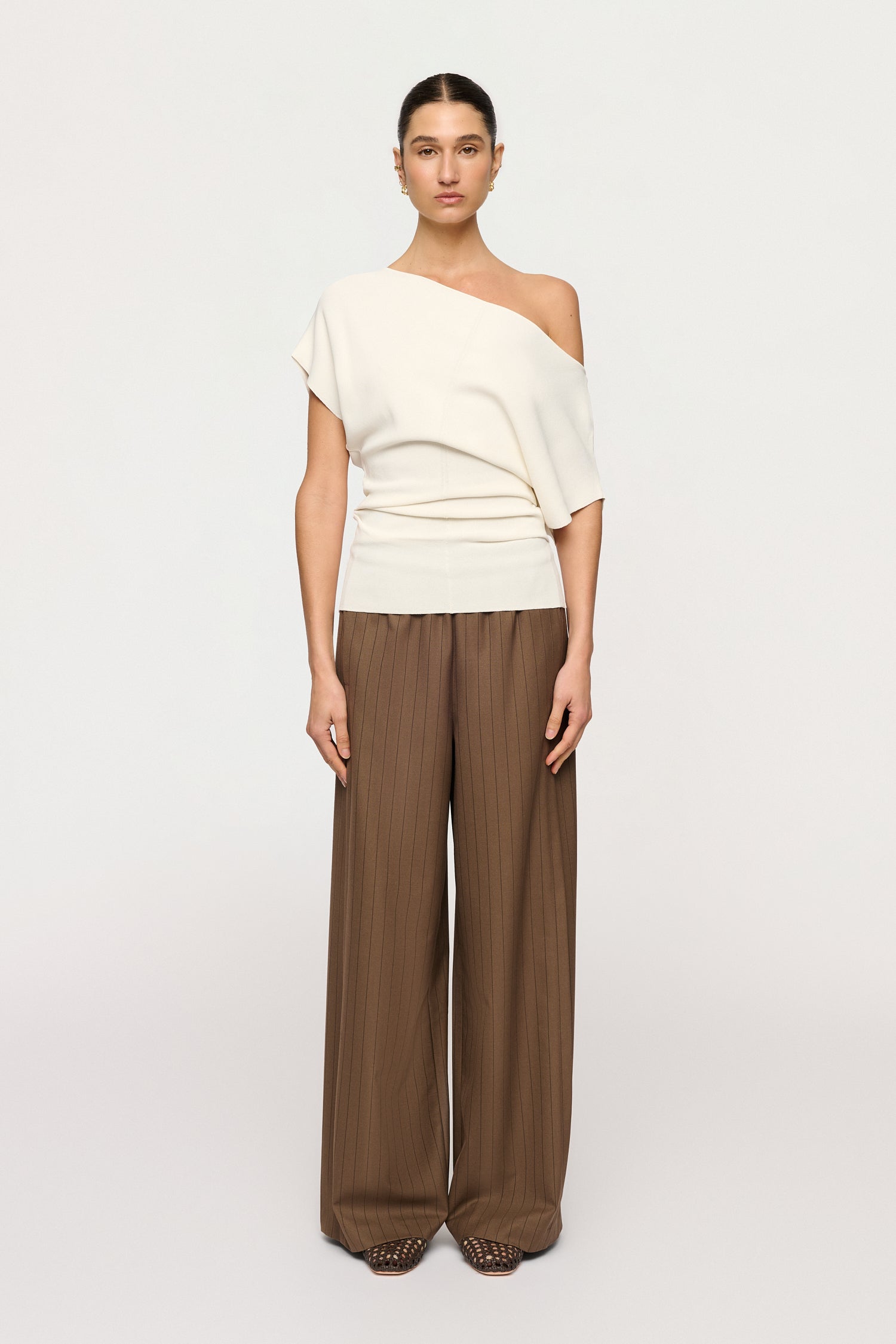 Woman wearing a white off-shoulder top and brown wide-leg pants on a light gray background